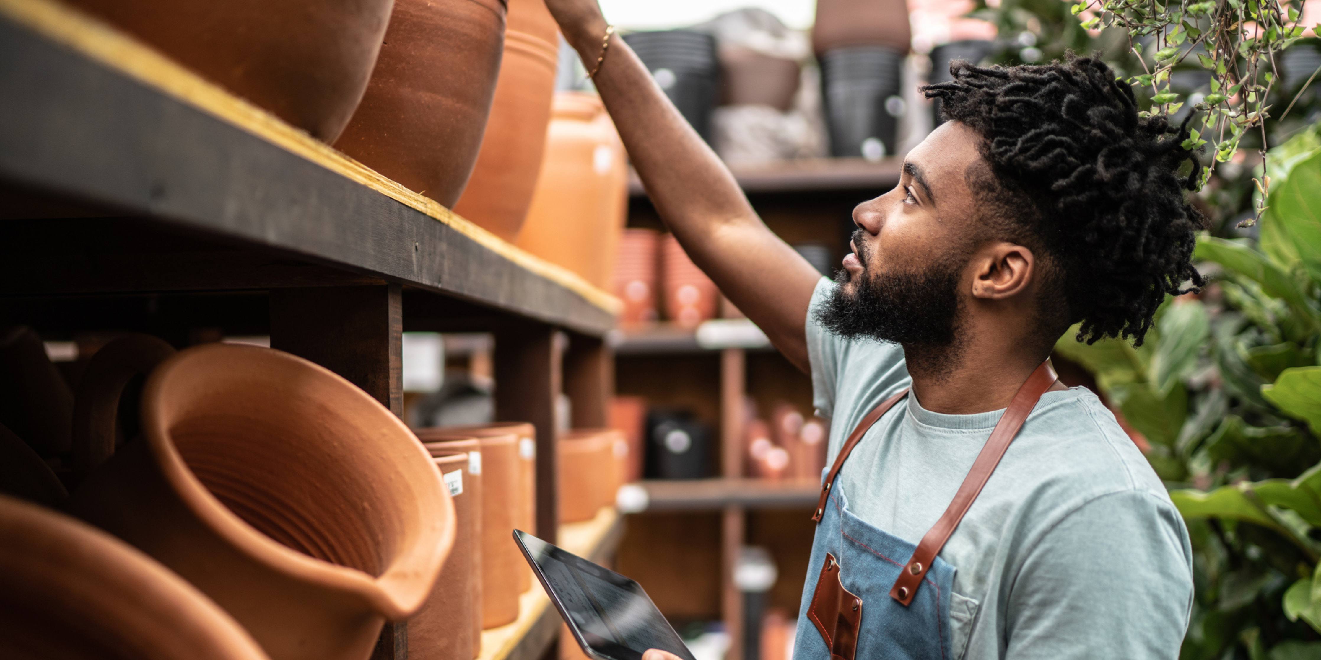Young salesman reaching overhead for a clay pot on a high shelf at a garden center