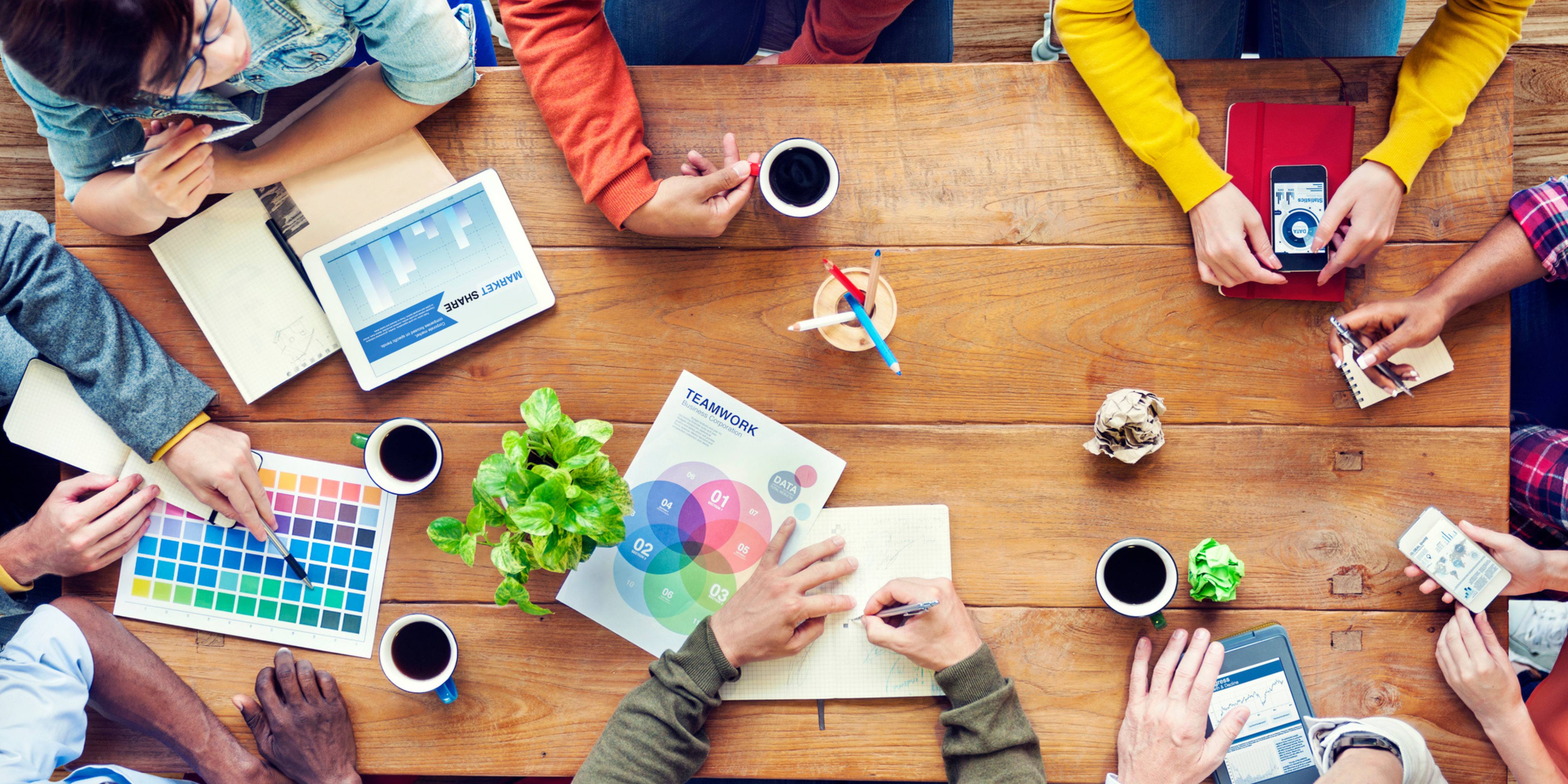 Group of people working together at a wooden table with various items like coffee cups, notebooks, and a tablet.