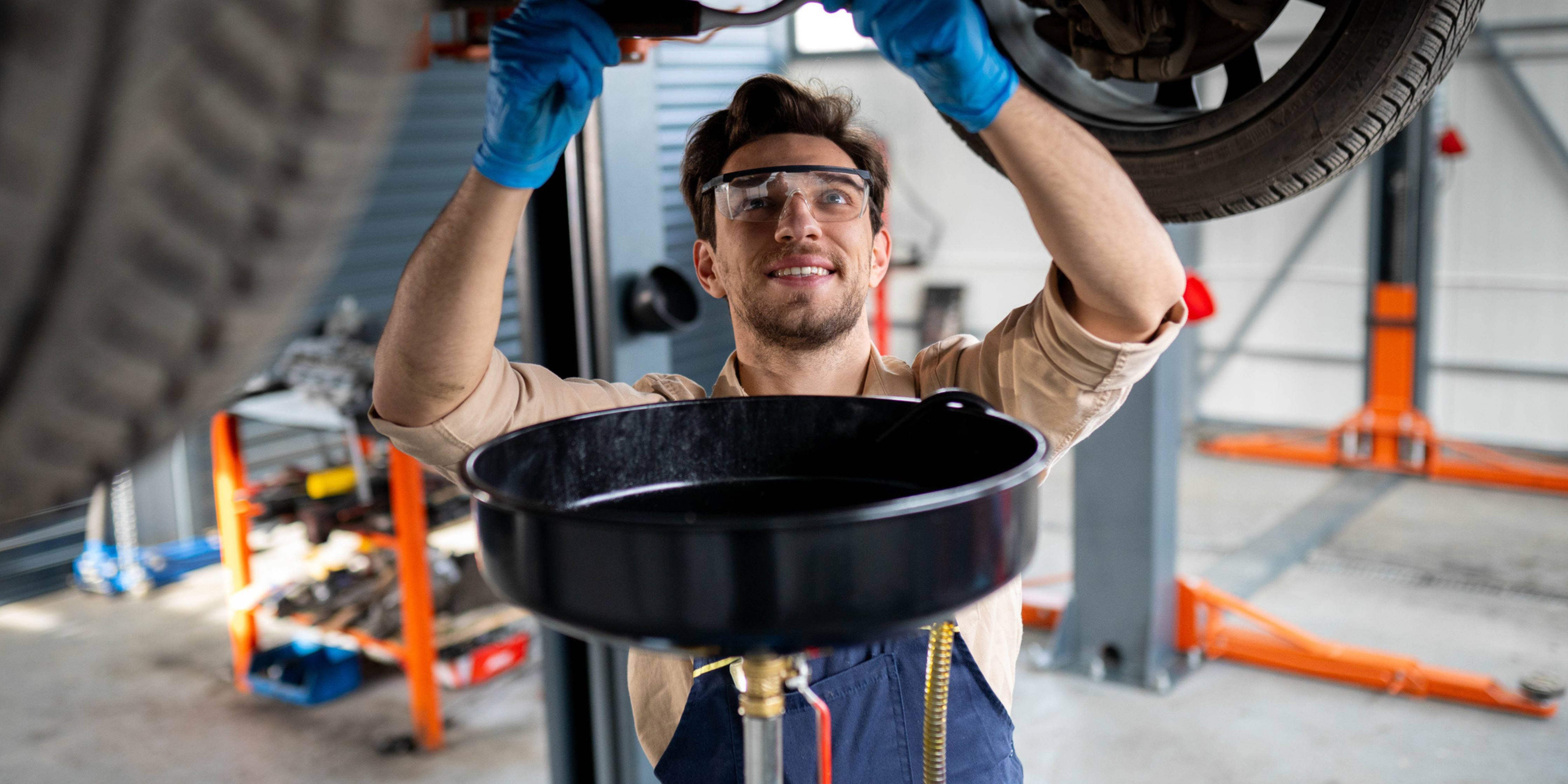 A mechanic is draining oil from the engine while standing under the car at mechanic's shop, wearing safety glasses and gloves.