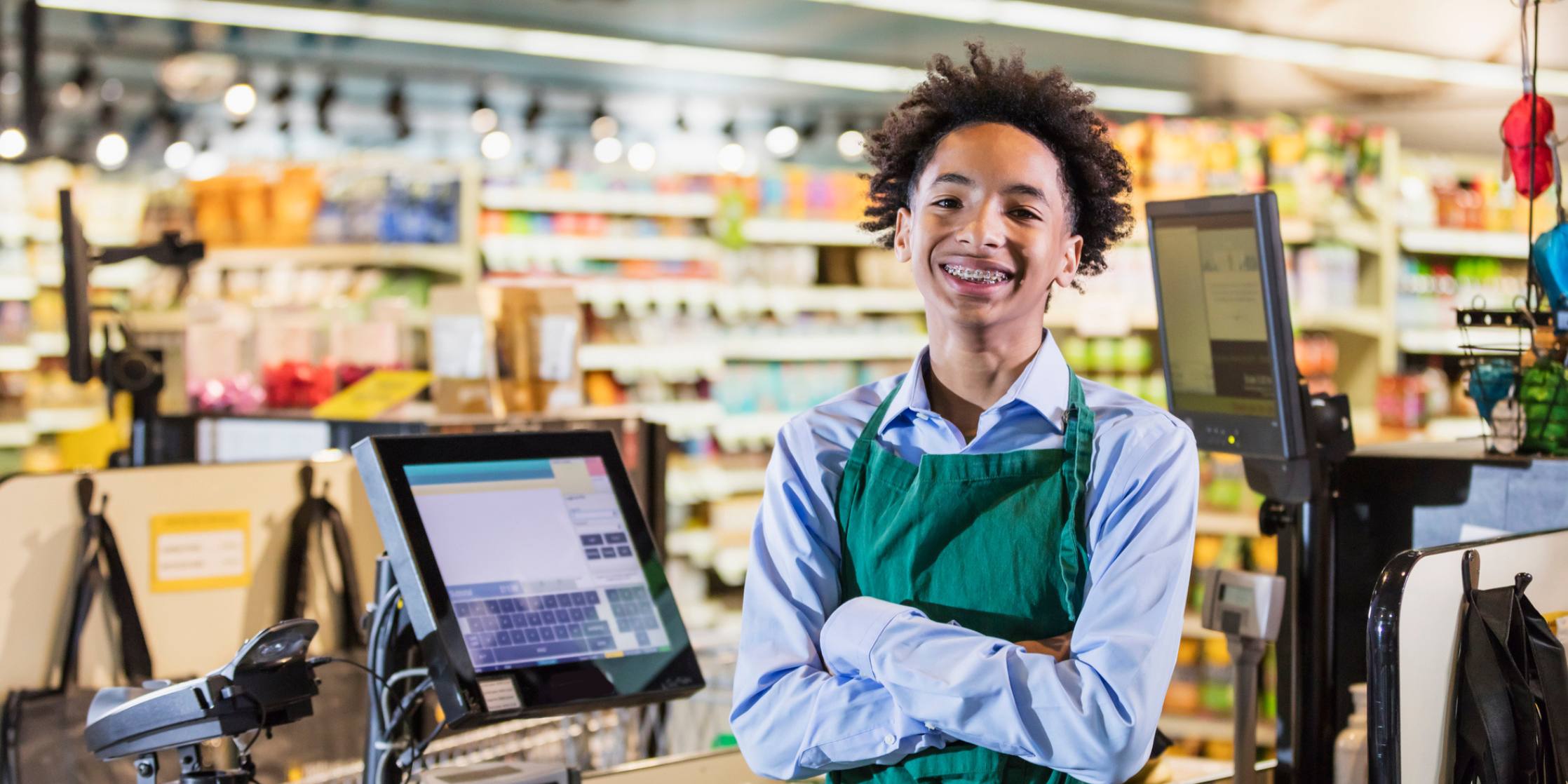 A mixed race African-American and Hispanic teenage boy working in a supermarket at the checkout counter. He is ready to scan groceries at the cash register. He is smiling at the camera.