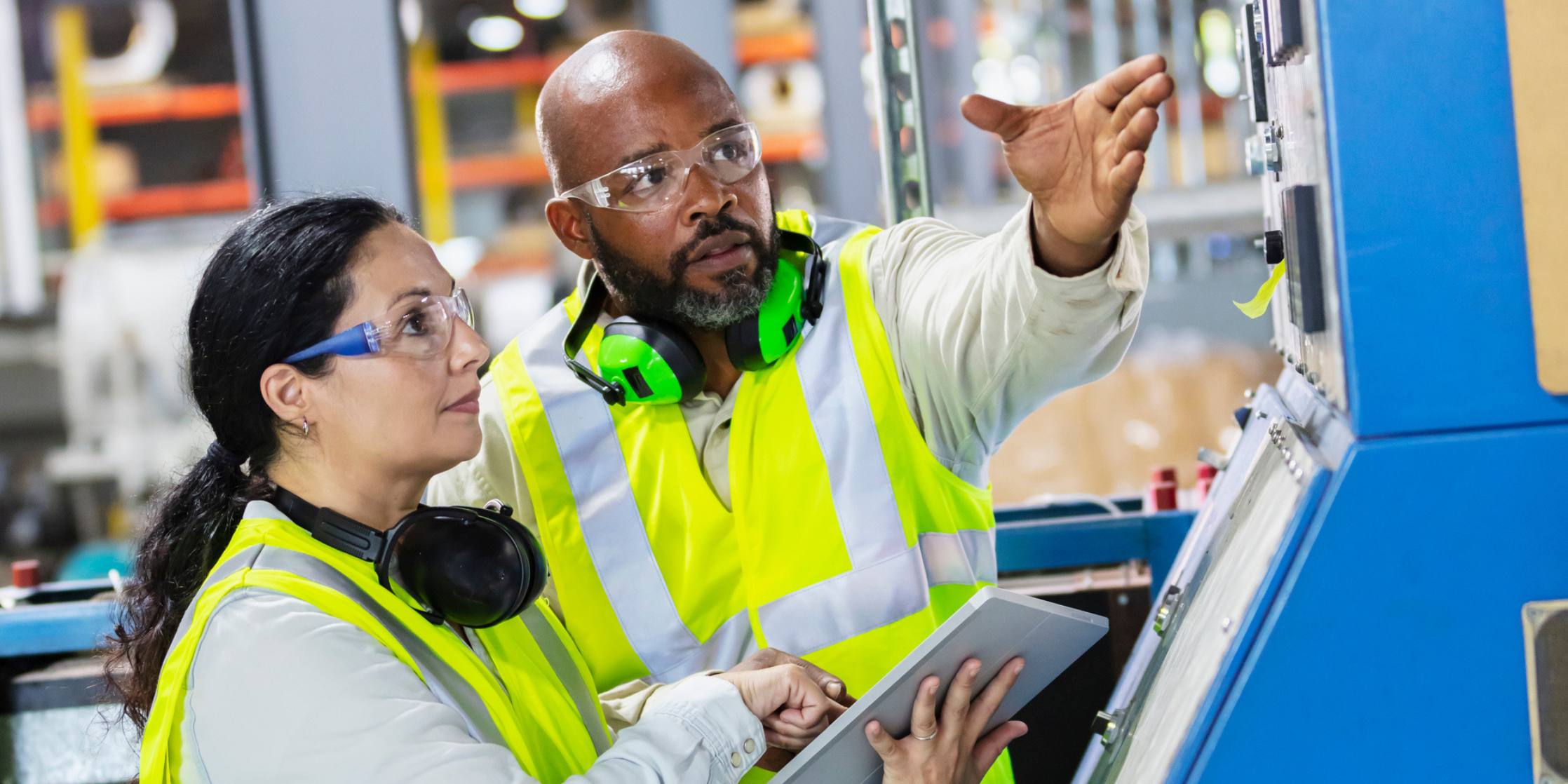 Two diverse factory workers reviewing a machine control panel; the woman holds a tablet and points to the screen while her male coworker gestures toward the panel.