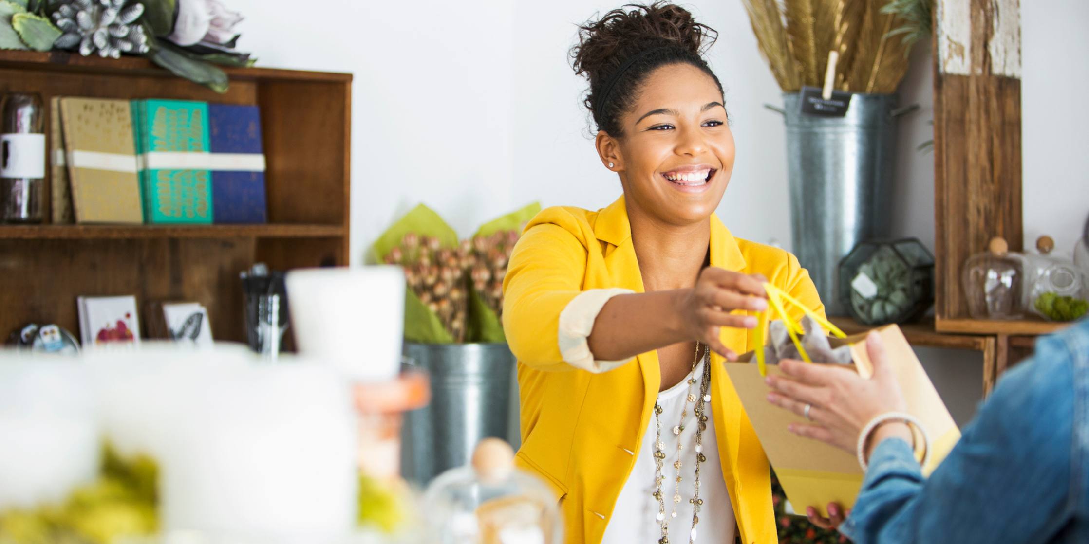 Woman in yellow jacket smiling and handing a shopping bag to a customer from behind a counter