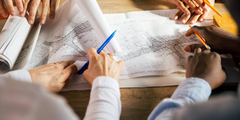 Over the shoulder view of two unrecognizable architects sitting in a meeting with their colleagues and reviewing technical drawings of a project they are designing.