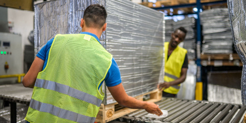 Diverse coworkers in an warehouse properly preparing a parcel for shipping. Wearing reflective clothes.