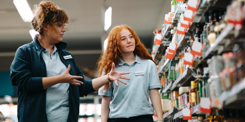 Two employees in a store aisle interacting with each other.