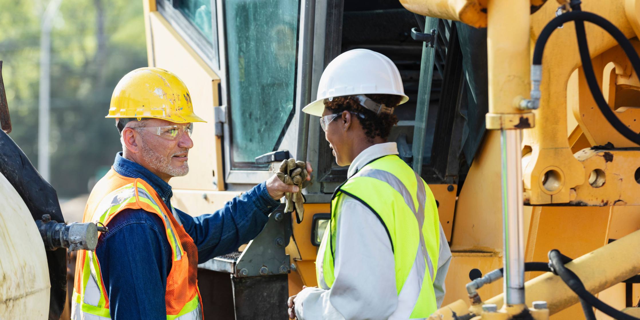 Two construction workers in front of a large piece of machinery on a construction site.