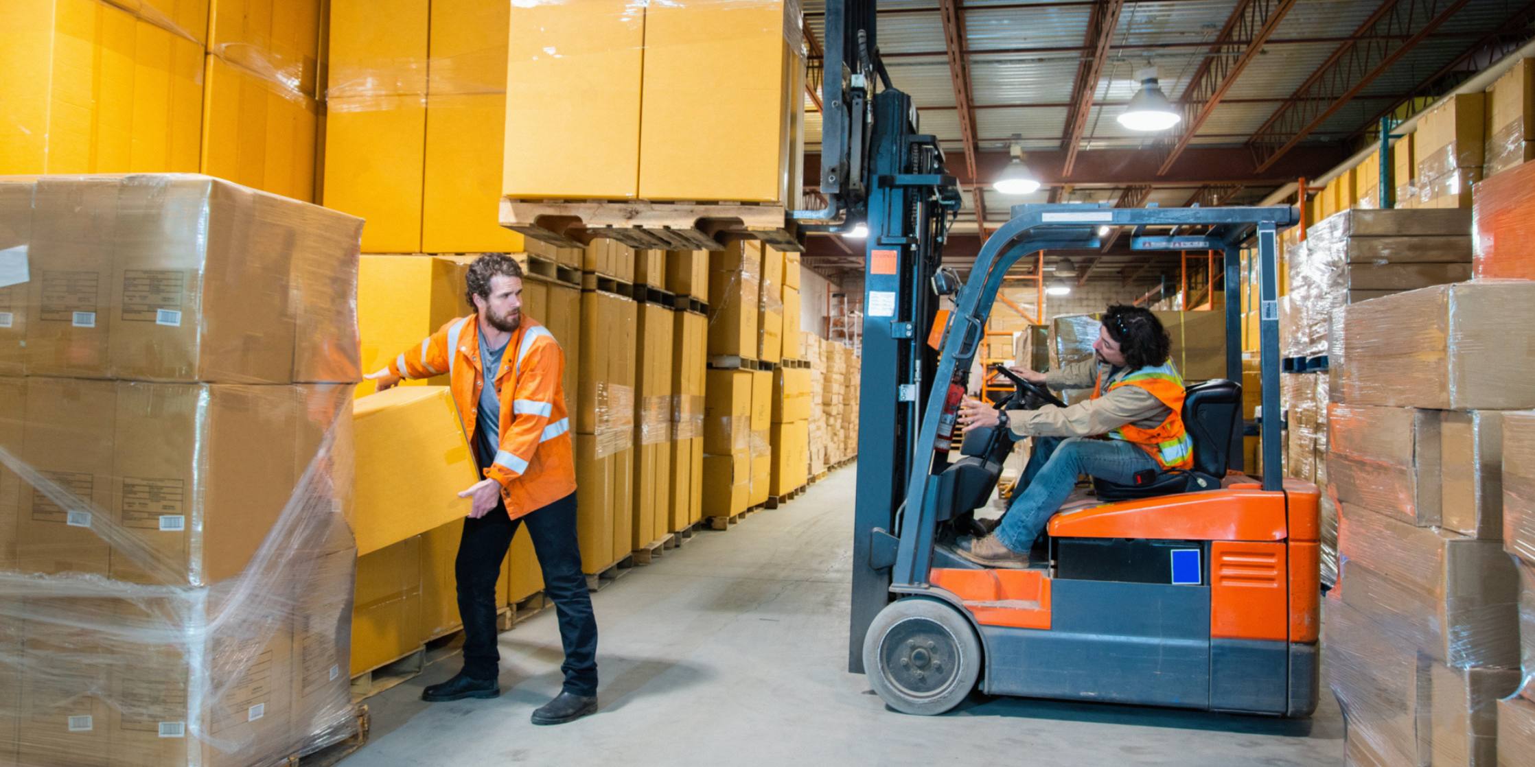 Two workers in a warehouse with forklift and stacked boxes