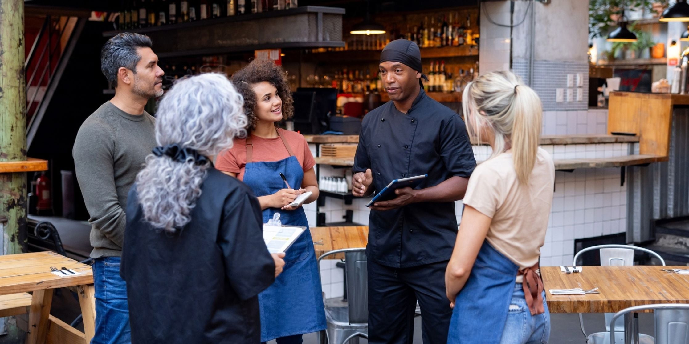 A group of five people, including two chefs in black uniforms and two servers in blue aprons, standing and talking in a restaurant setting.