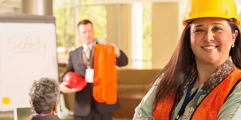 A smiling woman in a safety helmet and reflective vest with colleagues in the background in a training setting.